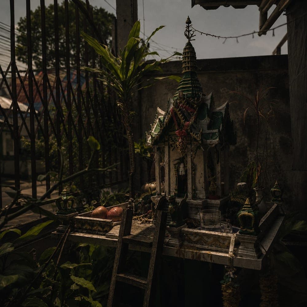 A Thai spirit house (ศาลพระภูมิ) with offerings of fruit and water among plants and an old wall, Chiang Mai.